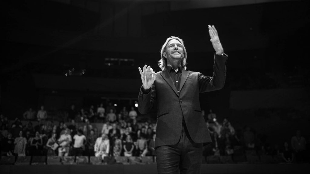Black and white dramatic photograph of conductor Eric Whitacre leading an orchestra and choir performance.