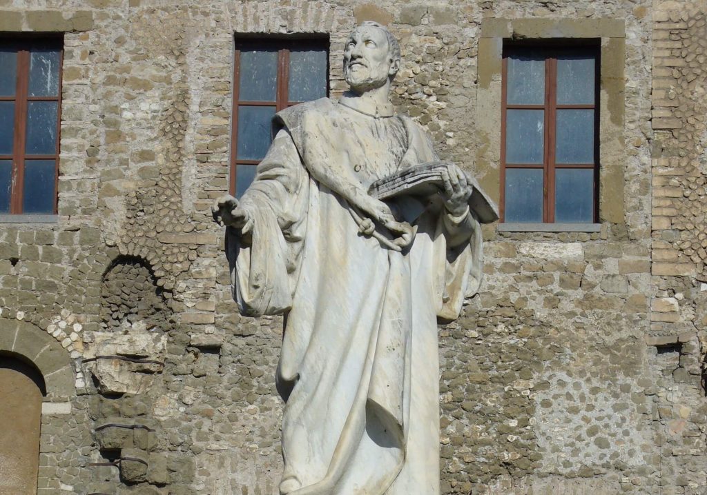 Full-length marble statue of Giovanni Pierluigi da Palestrina holding an open music book against a stone building in Italy.