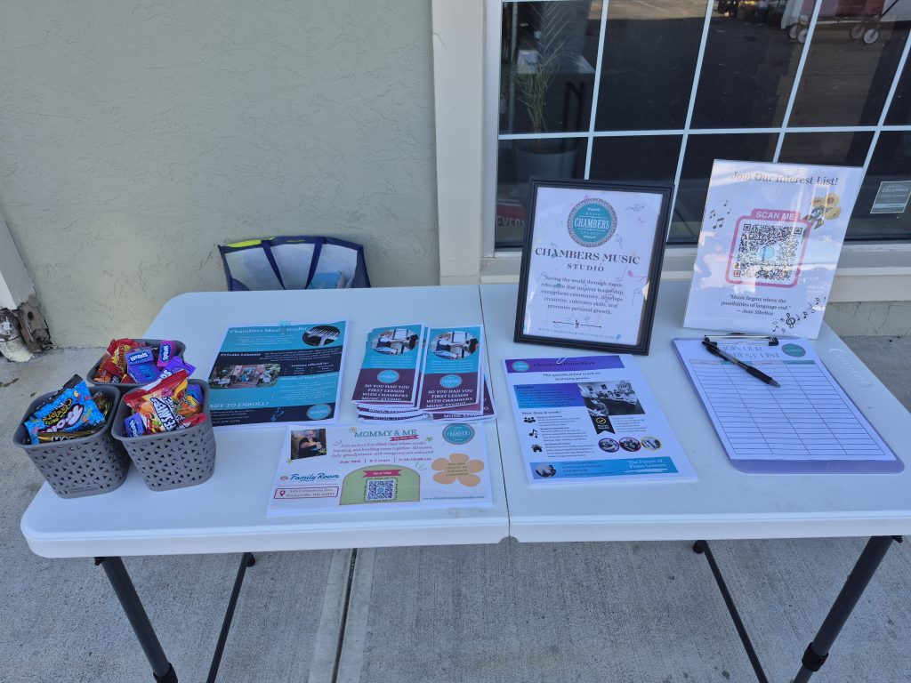 A white table displaying Chambers Music Studio program fliers, program interest sign-up sheets, and baskets of treats outside in Worthington.