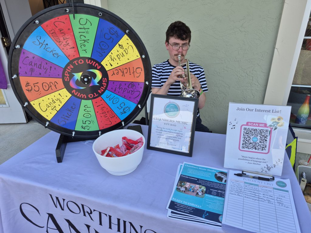 A Chambers Music Studio team member playing a trumpet behind an informational booth equipped with a prize wheel and QR code interest sheet.