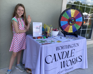 A young girl standing next to the Chambers Music Studio activity table outside Worthington Candle Works, featuring a colorful prize wheel and interest sign-up sheet.