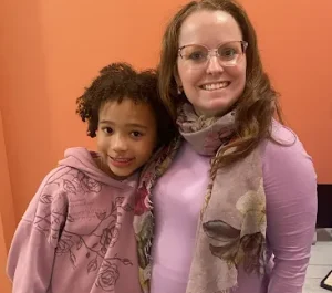 Julie and Eva standing together smiling indoors against an orange wall.