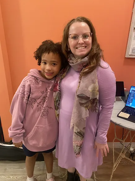 A close-up portrait of Julie in a light purple dress and Eva in a pink hoodie smiling at the camera.