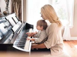 Home 47 image1 1 A mother gently guides her toddler’s hands to play a black upright piano in a bright, sunlit room, illustrating early childhood music education.