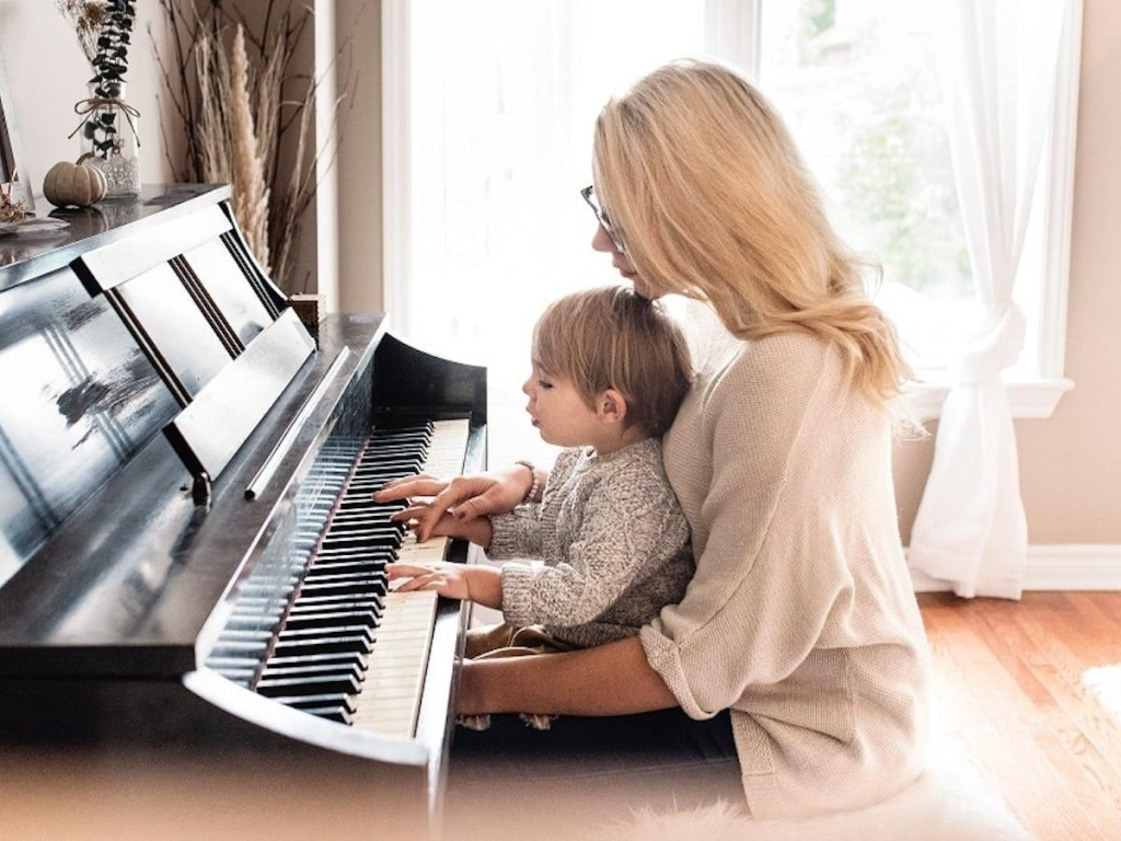 A mother sitting at an upright piano in a bright, sunlit room, guiding her toddler's hands as they place their fingers on the piano keys together.